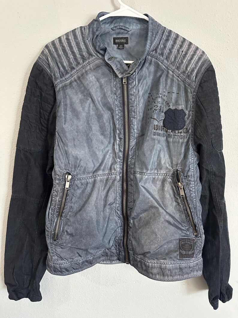 K&ouml;nnte beinhalten: Blau-schwarze Jacke mit Rei&szlig;verschluss. Die Jacke hat ein gestepptes Design an den Schultern und schwarze &Auml;rmel. Ein Aufn&auml;her mit Text und einem Schilddesign ist sichtbar. Die Jacke h&auml;ngt an einem wei&szlig;en Kleiderb&uuml;gel.