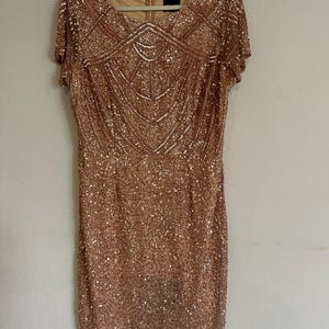 Puede incluir: Un vestido de lentejuelas en color oro rosa con mangas cortas, cuello redondo y un patr&oacute;n geom&eacute;trico de cuentas en el corpi&ntilde;o. La prenda est&aacute; completamente cubierta de lentejuelas brillantes e incluye un forro beige con una cremallera visible.