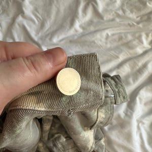 May include: A pair of light gray denim pants featuring a textured, distressed wash with subtle green and white mottled patterns. The waistband includes a silver-toned metal button embossed with the text MANIERE DE VOIR and a metal zipper fly.