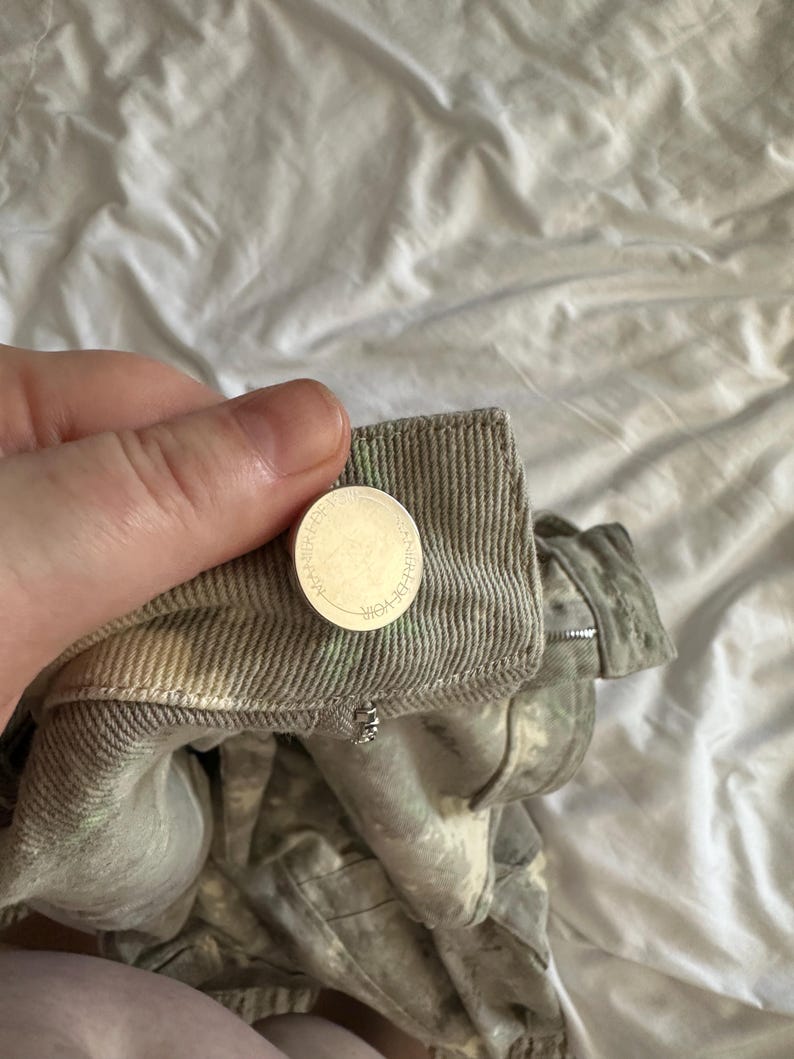 May include: A pair of light gray denim pants featuring a textured, distressed wash with subtle green and white mottled patterns. The waistband includes a silver-toned metal button embossed with the text MANIERE DE VOIR and a metal zipper fly.