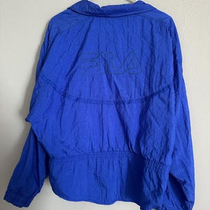 May include: A bright blue jacket featuring the FILA logo embroidered on the back. The jacket has a collared neckline, long sleeves, and an elasticated waist and cuffs. The jacket is hanging on a white hanger.