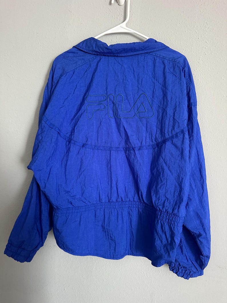 May include: A bright blue jacket featuring the FILA logo embroidered on the back. The jacket has a collared neckline, long sleeves, and an elasticated waist and cuffs. The jacket is hanging on a white hanger.
