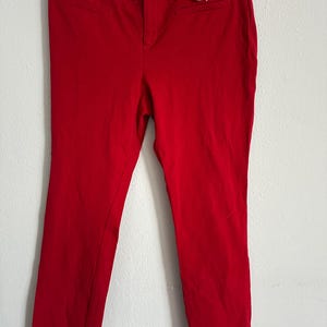 May include: Bright red pants with a button closure and pockets. The pants are hanging on a black hanger with a price tag attached. The pants appear to be made of a smooth, woven fabric.