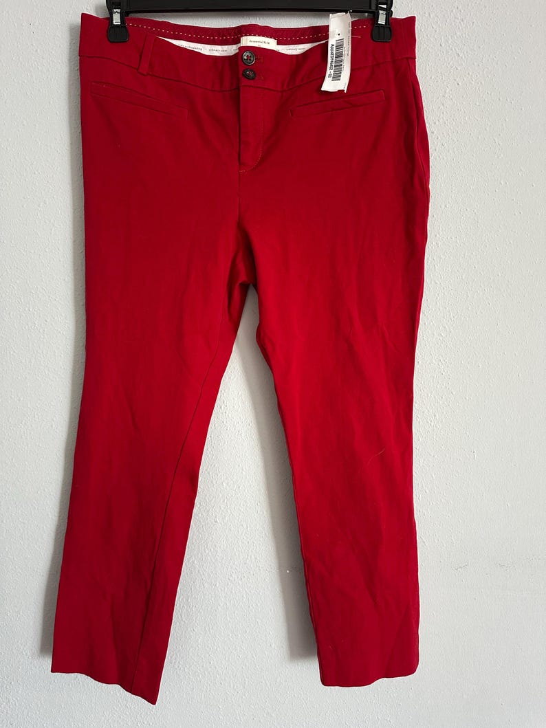 May include: Bright red pants with a button closure and pockets. The pants are hanging on a black hanger with a price tag attached. The pants appear to be made of a smooth, woven fabric.