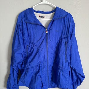 May include: A bright blue FILA jacket with a full zip and two side pockets. The jacket features the FILA logo on the chest and collar. It is suspended from a white hanger.