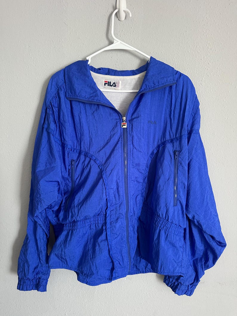 May include: A bright blue FILA jacket with a full zip and two side pockets. The jacket features the FILA logo on the chest and collar. It is suspended from a white hanger.