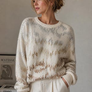 Pode incluir: Camisola creme com detalhes de lantejoulas prateadas e douradas. A camisola tem decote redondo e punhos canelados. A modelo usa calças brancas. Parte de uma coleção de roupas.