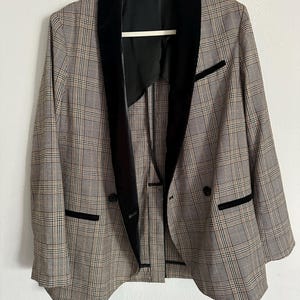 May include: A plaid blazer with a black velvet collar and trim. The jacket has a double-breasted design with two black buttons and two front pockets. The blazer is hanging on a white hanger against a white background.