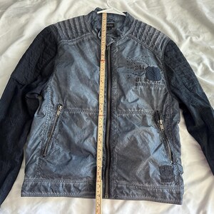K&ouml;nnte beinhalten: Eine blau-schwarze Jacke mit Rei&szlig;verschluss und Stehkragen. Die Jacke hat gesteppte Schulterpartien und schwarze &Auml;rmel. Auf der Vorderseite der Jacke befindet sich ein Aufn&auml;her mit Text und einem kleinen Emblem. Ein Ma&szlig;band ist vertikal auf der Jacke platziert.