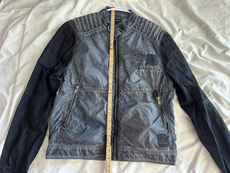 K&ouml;nnte beinhalten: Eine blau-schwarze Jacke mit Rei&szlig;verschluss und Stehkragen. Die Jacke hat gesteppte Schulterpartien und schwarze &Auml;rmel. Auf der Vorderseite der Jacke befindet sich ein Aufn&auml;her mit Text und einem kleinen Emblem. Ein Ma&szlig;band ist vertikal auf der Jacke platziert.