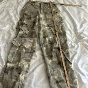 May include: Cargo pants featuring a mottled gray, white, and black camouflage-style pattern. The pants include two side cargo pockets with flaps and a waistband. A wooden folding ruler is placed across the waist and down the leg for scale.