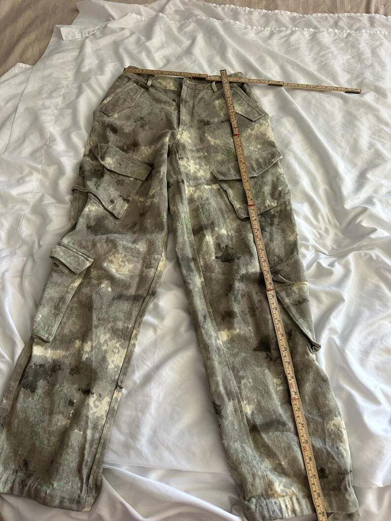 May include: Cargo pants featuring a mottled gray, white, and black camouflage-style pattern. The pants include two side cargo pockets with flaps and a waistband. A wooden folding ruler is placed across the waist and down the leg for scale.