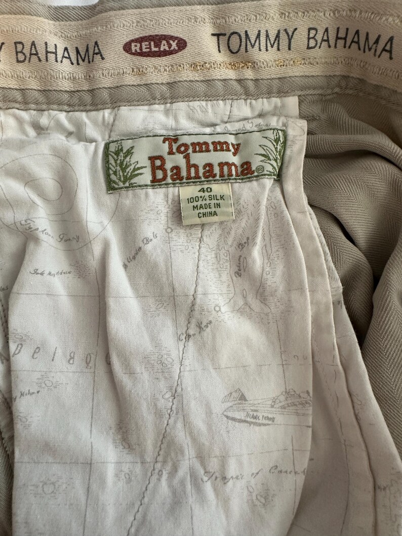 May include: Close-up of the interior of khaki-coloured Tommy Bahama trousers. The lining features a map design. The tag reads "Tommy Bahama" and "100% Silk". The waistband has the words "Tommy Bahama" and "Relax".