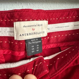 May include: Close-up of red pants with white stitching details. The label reads "the essential SLIM by ANTHROPOLOGIE" with size information: US 12, UK 16, EU 44. Made in The Philippines.
