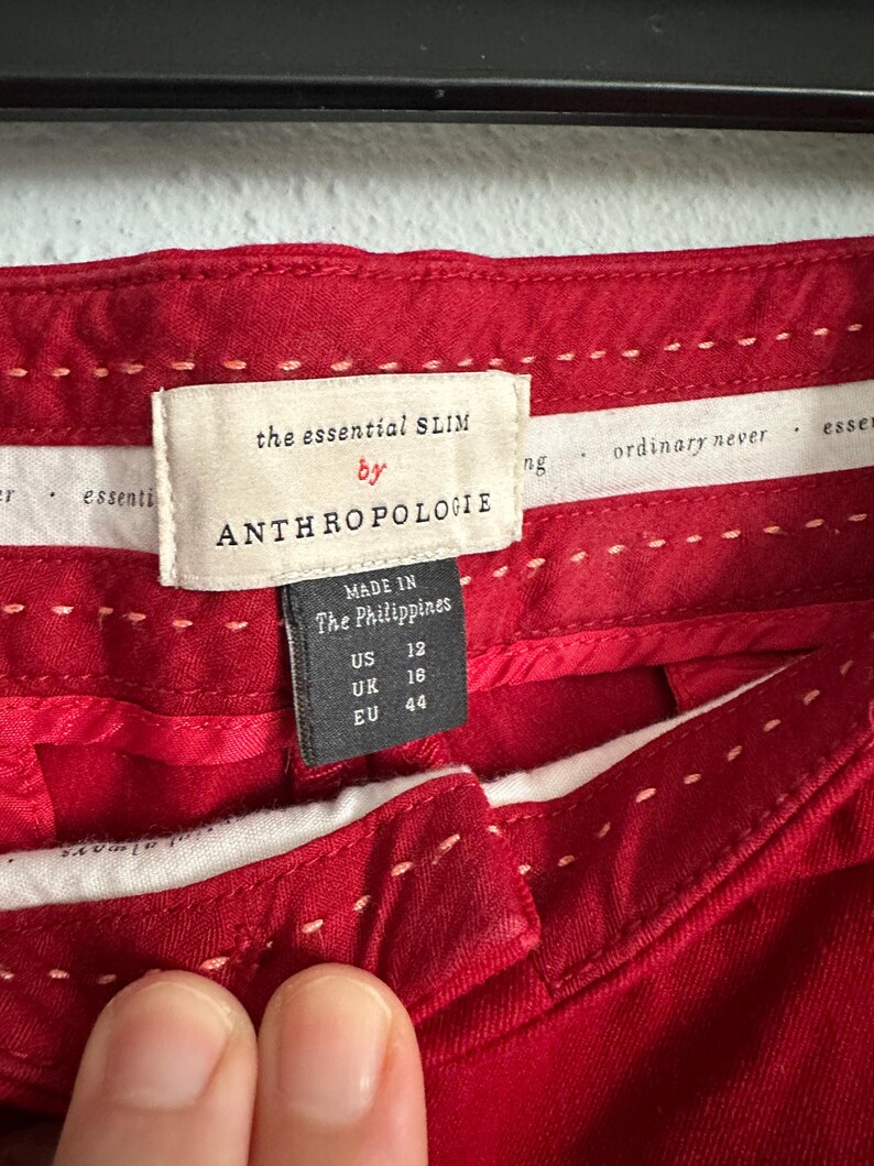 May include: Close-up of red pants with white stitching details. The label reads "the essential SLIM by ANTHROPOLOGIE" with size information: US 12, UK 16, EU 44. Made in The Philippines.