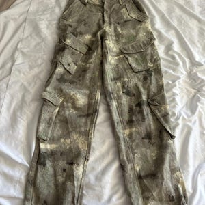 May include: Cargo pants featuring a mottled camouflage pattern in shades of olive green, brown, and cream. These pants include two large side cargo pockets with flaps, two front hip pockets, and adjustable ties at the bottom of each pant leg.