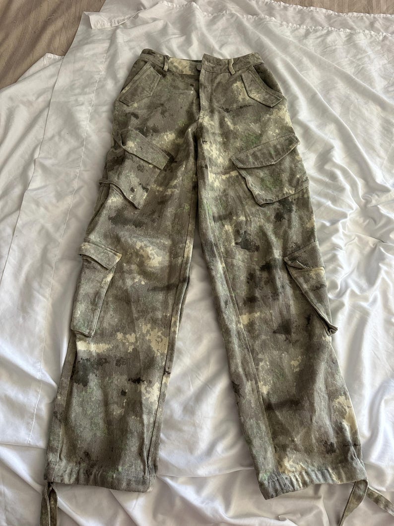 May include: Cargo pants featuring a mottled camouflage pattern in shades of olive green, brown, and cream. These pants include two large side cargo pockets with flaps, two front hip pockets, and adjustable ties at the bottom of each pant leg.