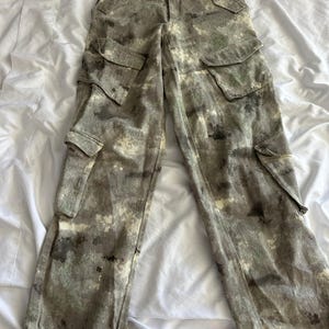 May include: High-waisted cargo pants featuring a mottled, camouflage-like pattern in shades of olive green, charcoal gray, and off-white. These pants include two large side cargo pockets, two hip pockets, and adjustable drawstring ties at the leg hems.