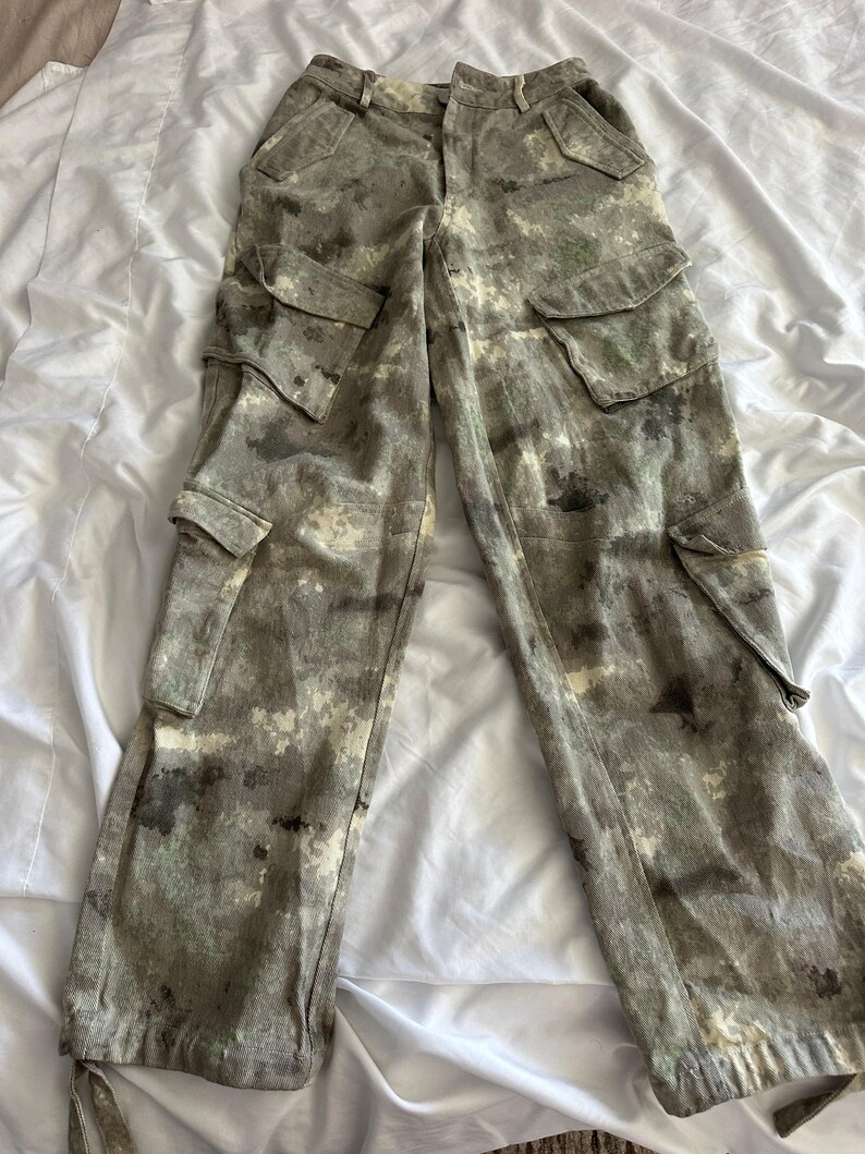 May include: High-waisted cargo pants featuring a mottled, camouflage-like pattern in shades of olive green, charcoal gray, and off-white. These pants include two large side cargo pockets, two hip pockets, and adjustable drawstring ties at the leg hems.