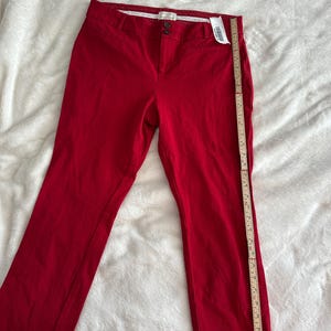 May include: Bright red pants with a straight leg cut. The pants have a button and zipper closure, belt loops, and pockets. A measuring tape is placed next to the pants. The pants are made of a smooth fabric.