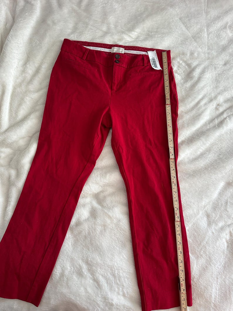 May include: Bright red pants with a straight leg cut. The pants have a button and zipper closure, belt loops, and pockets. A measuring tape is placed next to the pants. The pants are made of a smooth fabric.
