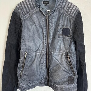 K&ouml;nnte beinhalten: Grau-schwarze Jacke mit Rei&szlig;verschluss vorne und zwei Rei&szlig;verschlusstaschen. Die Jacke hat einen Used-Look mit gesteppten Schulterdetails und schwarzen &Auml;rmeln. Ein Aufn&auml;her auf der Brust zeigt "Buffalo David Bitton".