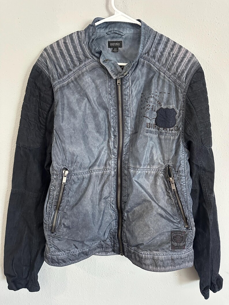 K&ouml;nnte beinhalten: Grau-schwarze Jacke mit Rei&szlig;verschluss vorne und zwei Rei&szlig;verschlusstaschen. Die Jacke hat einen Used-Look mit gesteppten Schulterdetails und schwarzen &Auml;rmeln. Ein Aufn&auml;her auf der Brust zeigt "Buffalo David Bitton".