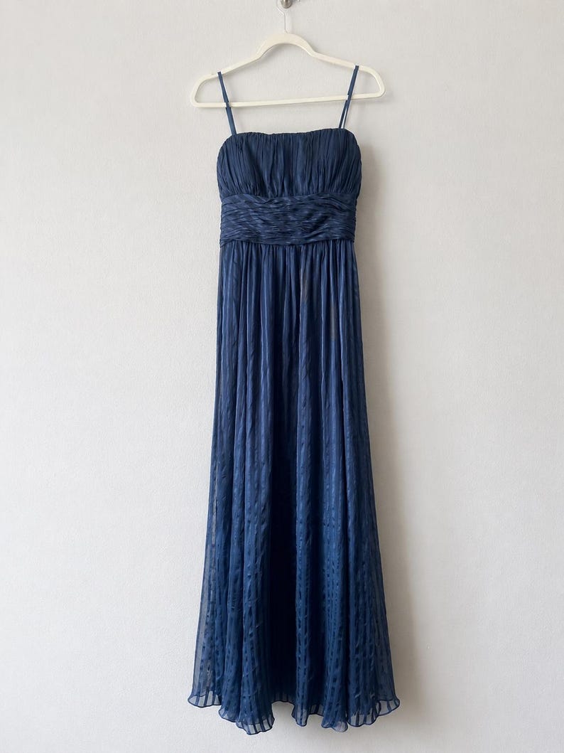 Peut inclure: Une robe de soir&eacute;e bleu marine avec des bretelles fines, un corsage fronc&eacute; et une taille ajust&eacute;e. La jupe longue pr&eacute;sente un motif subtil. La robe est suspendue &agrave; un cintre blanc sur un fond uni.