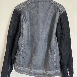 K&ouml;nnte beinhalten: Grau-schwarze Jacke mit verwaschenem, strukturiertem Aussehen. Die Jacke hat einen Kragen, gesteppte Schulterdetails und schwarze &Auml;rmel mit Rei&szlig;verschlussmanschetten. Die R&uuml;ckseite der Jacke ist hellgrau und wirkt dezent abgenutzt.