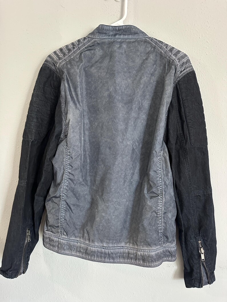 K&ouml;nnte beinhalten: Grau-schwarze Jacke mit verwaschenem, strukturiertem Aussehen. Die Jacke hat einen Kragen, gesteppte Schulterdetails und schwarze &Auml;rmel mit Rei&szlig;verschlussmanschetten. Die R&uuml;ckseite der Jacke ist hellgrau und wirkt dezent abgenutzt.