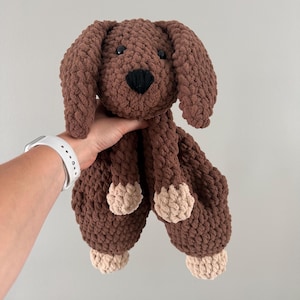 May include: A handmade brown crochet dog plushie with a black nose and small black eyes. The dog has long floppy ears and tan paws. The plush toy is soft and cuddly, perfect for children.