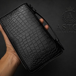 Könnte beinhalten: Schwarze Ledergeldbörse mit Krokodilleder-Textur. Die Geldbörse hat einen Reißverschluss und eine Handschlaufe. Das Astor Leather-Logo ist sichtbar. Die Geldbörse wird in einer Hand gehalten.