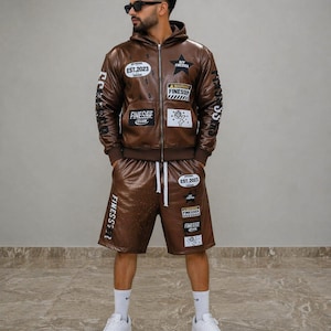 May include: A brown faux leather hoodie and shorts set. The jacket has patches with text and graphics, including "EST.2023" and "OFF FINESSER". The shorts also have patches and a drawstring. The person is wearing white trainers.