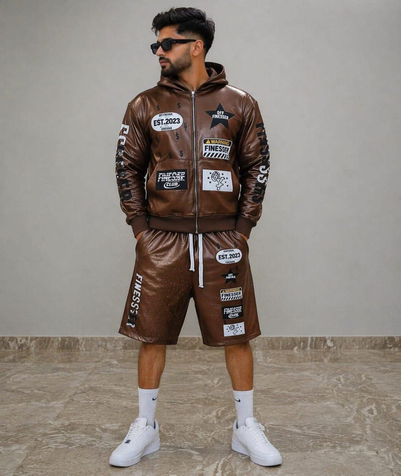 May include: A brown faux leather hoodie and shorts set. The jacket has patches with text and graphics, including "EST.2023" and "OFF FINESSER". The shorts also have patches and a drawstring. The person is wearing white trainers.