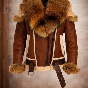 Könnte beinhalten: Braune Lederjacke mit großem Pelzkragen und Manschetten. Die Jacke hat einen Gürtel in der Taille, einen Reißverschluss und hellfarbene Verzierungen. Das Fell ist eine Mischung aus Braun- und Beigetönen.