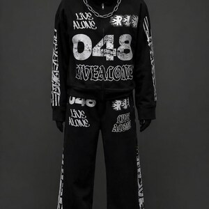 May include: Black hooded sweatshirt and matching sweatpants set. The sweatshirt features the text "LIVE ALONE" and "048 LIVEALONE" in white. A silver chain necklace is worn with the hoodie. The sweatpants also have white text. Black sneakers complete the outfit.