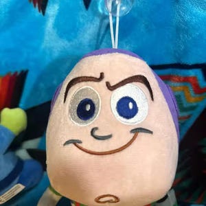 May include: A plush Buzz Lightyear toy with a smiling face, purple head, and green and white spacesuit. The toy has a suction cup attached to the top. The background is a blue and multicolored patterned fabric.