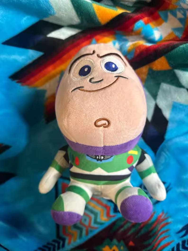 May include: A plush Buzz Lightyear doll, featuring a light pink head with embroidered facial details, including blue eyes and a smiling expression. The doll is dressed in a green, white, and purple spacesuit, with white gloves and purple boots. The background is a patterned blanket.