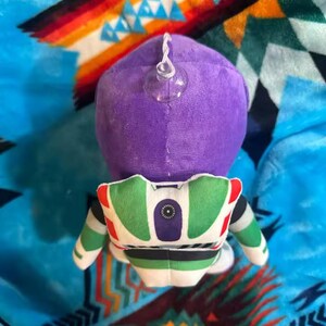 May include: A plush toy of Buzz Lightyear from Toy Story, viewed from the back. The toy features a purple head, a green and white spacesuit, and a clear suction cup attached to the head. The toy is on a colorful patterned blanket.