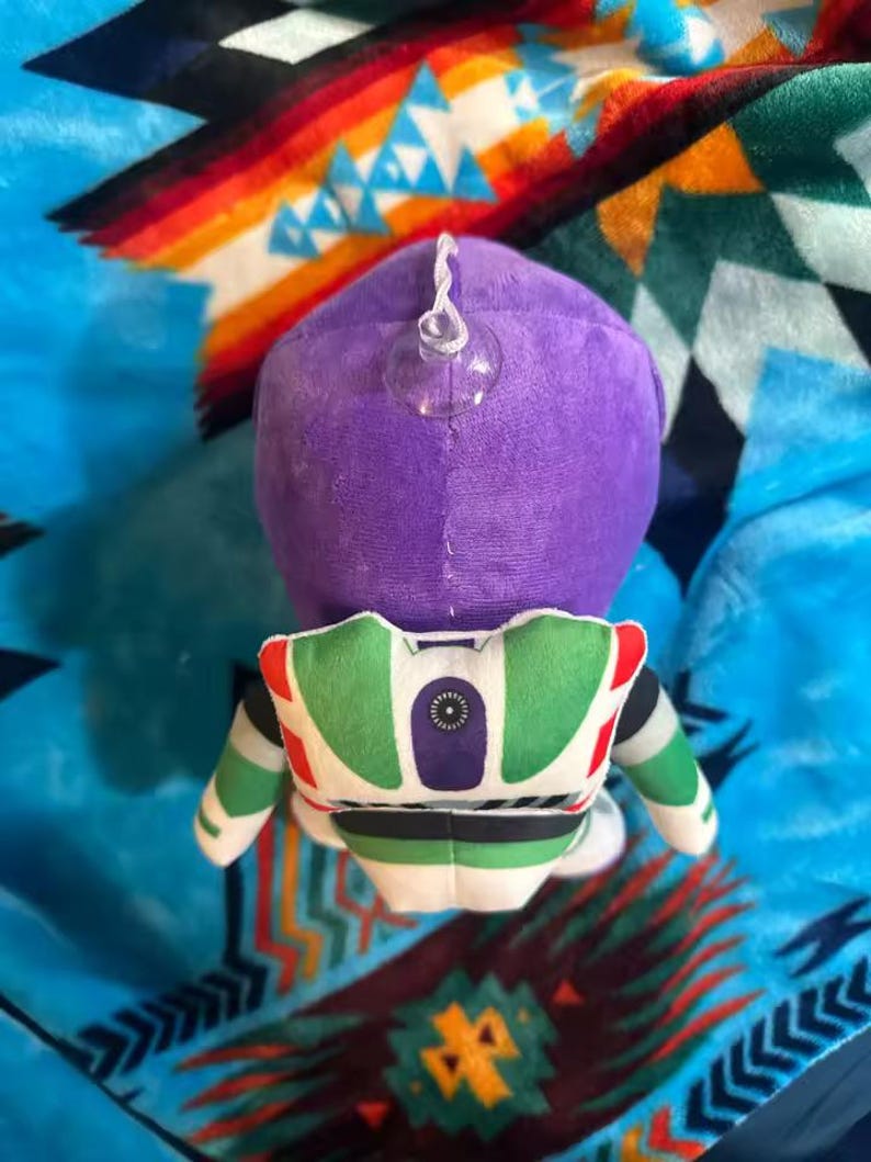May include: A plush toy of Buzz Lightyear from Toy Story, viewed from the back. The toy features a purple head, a green and white spacesuit, and a clear suction cup attached to the head. The toy is on a colorful patterned blanket.
