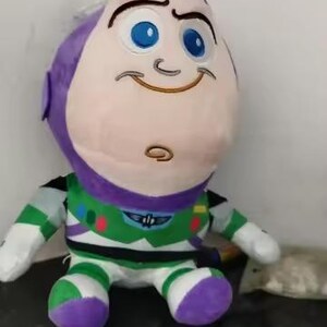 May include: A plush Buzz Lightyear doll, featuring a large head with a purple cap, blue eyes, and a smiling expression. The doll wears a green, white, and purple spacesuit. A fun toy for kids.