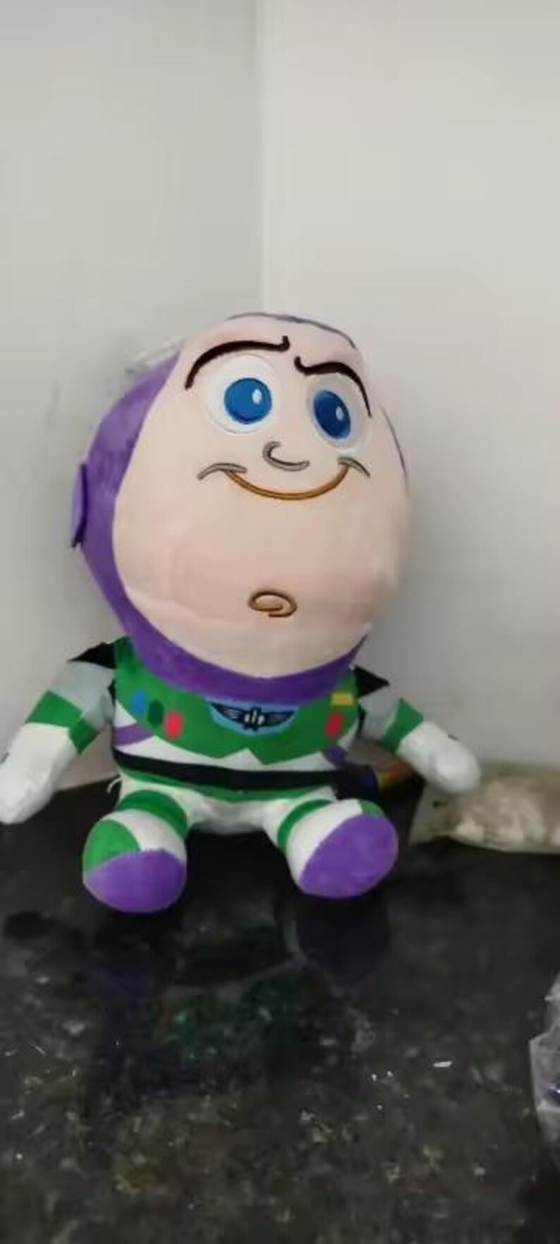 May include: A plush Buzz Lightyear doll, featuring a large head with a purple cap, blue eyes, and a smiling expression. The doll wears a green, white, and purple spacesuit. A fun toy for kids.