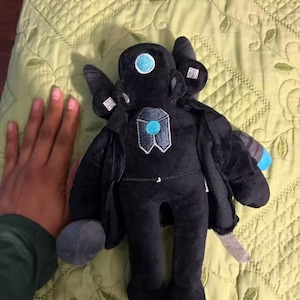 May include: A black plush toy with a unique design, featuring a round head with a blue circle, and a geometric emblem on the chest. The toy has a cape and a gray, rounded object attached to its side. The toy is approximately 8 inches tall.