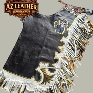 May include: Black leather chaps featuring white and gold decorative trim, long layered fringe in white and metallic gold, and an embroidered patch of a skeleton wearing a suit and hat. The upper section includes brown embossed leather detailing.