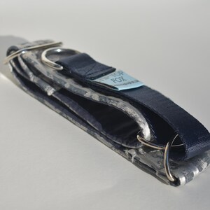 May include: A folded dog collar featuring a grey and white camouflage pattern on one side and a solid dark navy blue strap on the other. It includes silver-toned metal D-rings and a light blue fabric tag with the text TIP TOP FOX printed on it.
