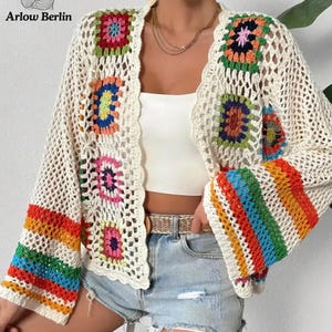 May include: A cream-colored open-front crochet cardigan featuring multicolored granny square motifs along the front panels and wide sleeves with horizontal rainbow stripes. The garment has a scalloped edge and an open-knit texture.