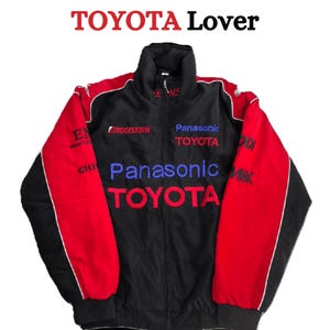 Può includere: Giacca da corsa Toyota nera e rossa con dettagli bianchi. La giacca presenta il testo "TOYOTA Lover" in alto e "Panasonic TOYOTA" in blu e rosso sul davanti. Le maniche sono rosse con dettagli neri e bianchi.