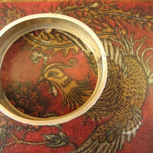 May include: A gold-colored hinged bangle bracelet. The bracelet is circular and has a clasp. The background is a red surface with a gold and brown phoenix design.