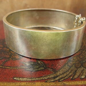 May include: A wide, silver-toned bangle bracelet with a simple, minimalist design. The bracelet has a smooth, brushed metal finish and a hinged clasp with a safety chain. The bracelet is approximately 2.5 inches in diameter.