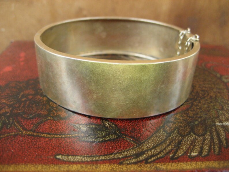 May include: A wide, silver-toned bangle bracelet with a simple, minimalist design. The bracelet has a smooth, brushed metal finish and a hinged clasp with a safety chain. The bracelet is approximately 2.5 inches in diameter.